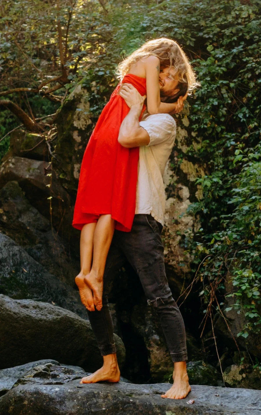 Rytis lifting Violeta in a red dress beside a forest waterfall