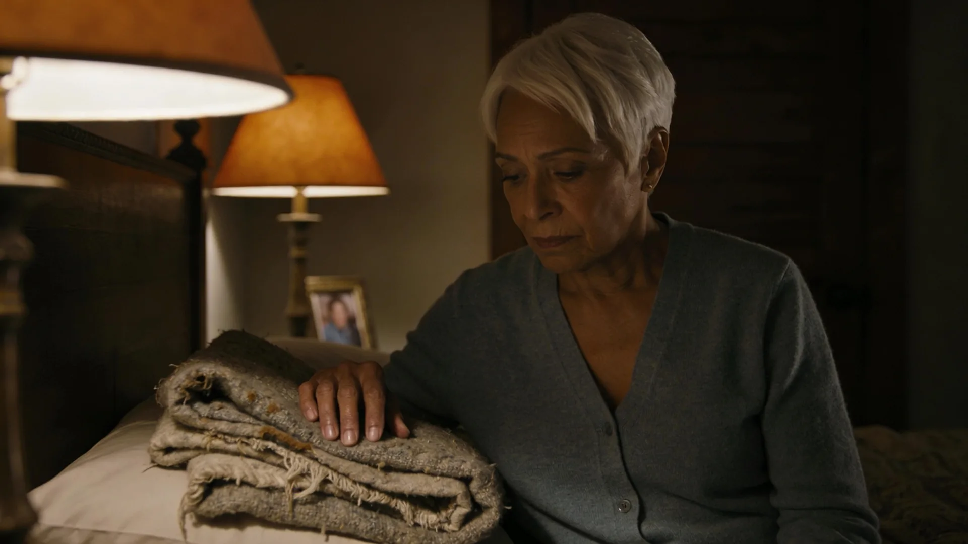 Grief ambush when trying to sleep — close-up of a hand on a folded woolen blanket beside a pillow in warm bedside lamp light