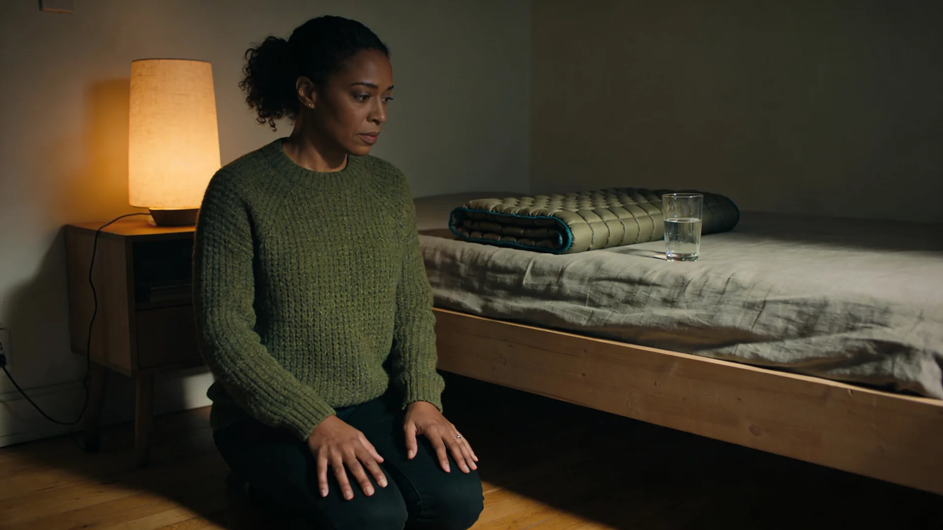 How to help someone having a panic attack — kneeling beside a bed where a weighted blanket and water sit ready, hands palm-down on thighs, helping someone 