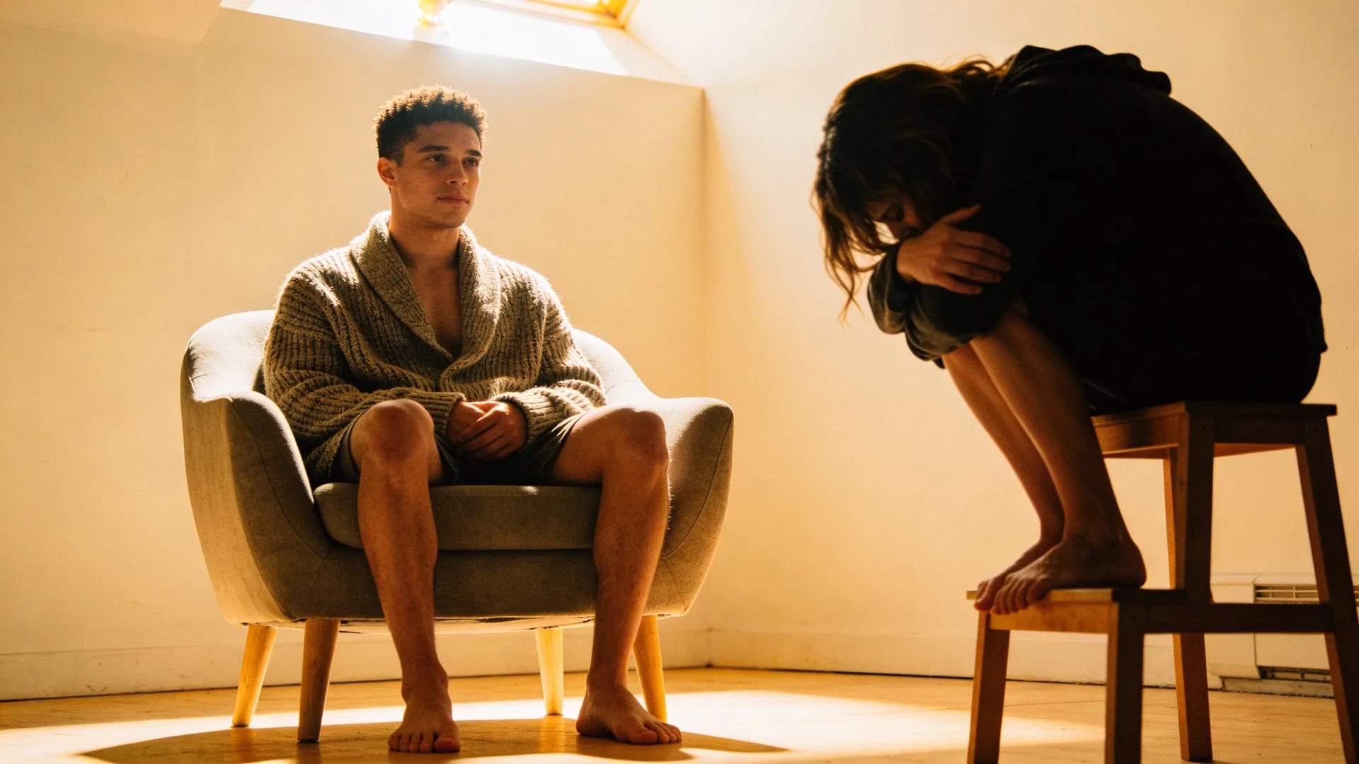 Woman hunched on a wooden stool in dim bathroom light, shoulders curved inward where repressed emotions settle in the body